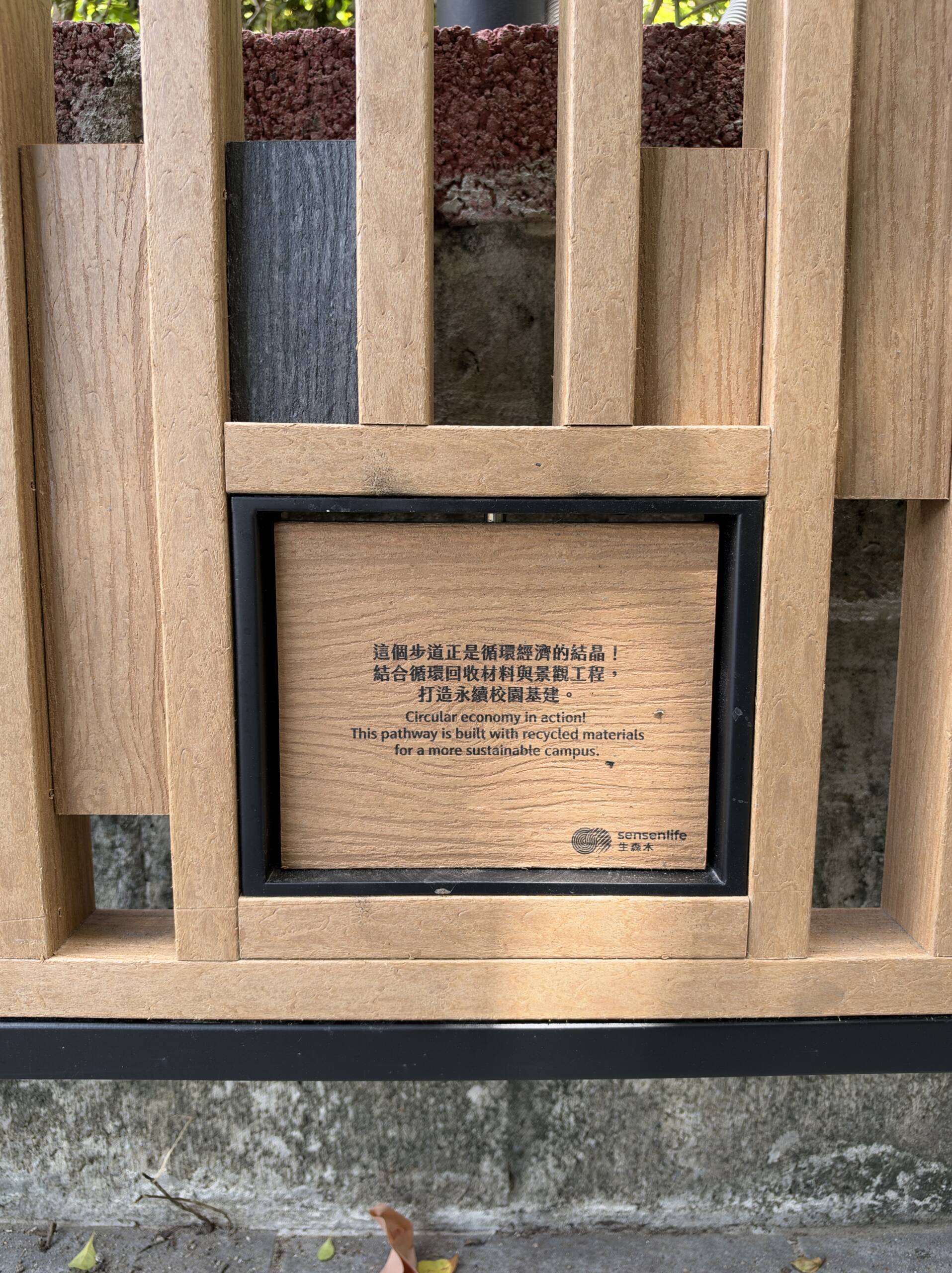 永續步道再生塑料木紋格柵上的生森木標示牌,中英文寫著步道以循環回收材料打造永續校園 永續步道再生塑料木紋格柵上的生森木標示牌,中英文寫著步道以循環回收材料打造永續校園